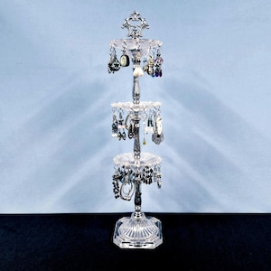 May include: A three-tiered silver and clear glass jewelry stand featuring a central vertical pole with decorative metal accents. Each tier has a circular glass tray with small holes along the edge for hanging various pairs of earrings.