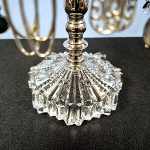 May include: A jewelry stand featuring a clear, faceted glass base with a starburst pattern and a central silver-toned metal post. The stand supports various hanging accessories, including gold-toned hoops and beaded ornaments with green and black accents.
