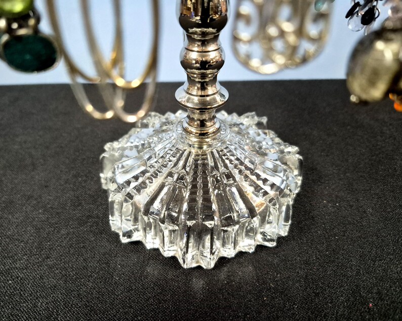May include: A jewelry stand featuring a clear, faceted glass base with a starburst pattern and a central silver-toned metal post. The stand supports various hanging accessories, including gold-toned hoops and beaded ornaments with green and black accents.