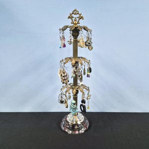 May include: A tall, tiered brass jewelry stand featuring three rotating levels with hooks for hanging earrings. The central pole is topped with an ornate finial, and the base is constructed from clear, faceted glass, providing a stable foundation.