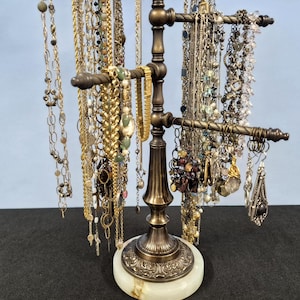 May include: A bronze-toned metal jewelry stand with multiple horizontal arms for hanging necklaces and bracelets. The stand features a decorative, tiered base set upon a circular, light-colored stone pedestal with a smooth, polished finish.