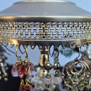 May include: An antique-style earring holder with a silver and gold finish. The holder features a decorative filigree design and is adorned with various earrings in shades of silver, pink, and gold. The earrings include beaded and crystal designs.