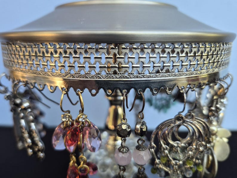 May include: An antique-style earring holder with a silver and gold finish. The holder features a decorative filigree design and is adorned with various earrings in shades of silver, pink, and gold. The earrings include beaded and crystal designs.