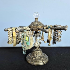 May include: A rotating jewelry display stand featuring an antique bronze-toned metal base with ornate floral detailing, a central clear faceted crystal pillar, and multiple horizontal arms holding various bracelets and metal wristwatches.