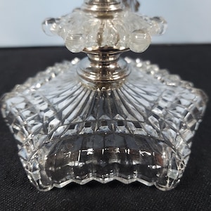 Pu&ograve; includere: Primo piano di una base di candelabro in cristallo trasparente e argento. La base ha un design quadrato a forma di diamante con un bordo smerlato. Lo stelo argentato supporta una parte superiore in cristallo trasparente con accenti arrotondati.