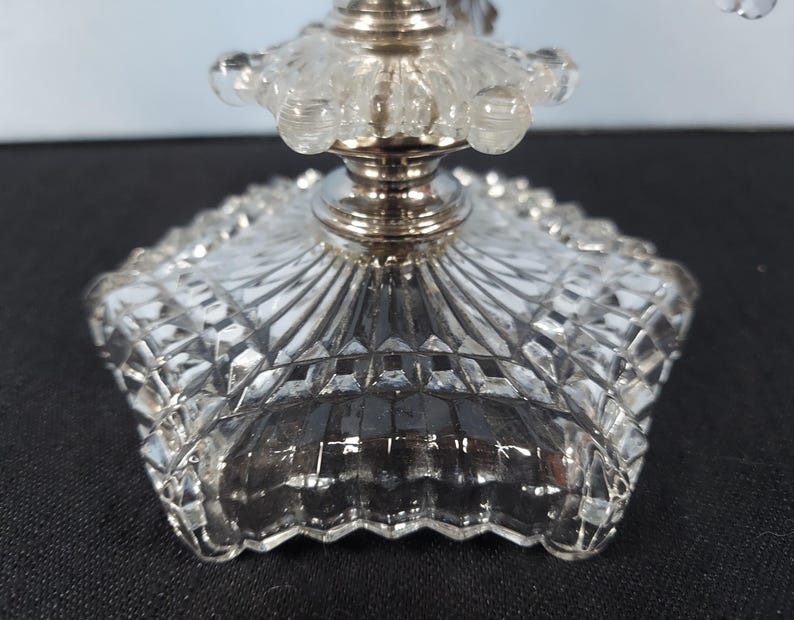 Pu&ograve; includere: Primo piano di una base di candelabro in cristallo trasparente e argento. La base ha un design quadrato a forma di diamante con un bordo smerlato. Lo stelo argentato supporta una parte superiore in cristallo trasparente con accenti arrotondati.