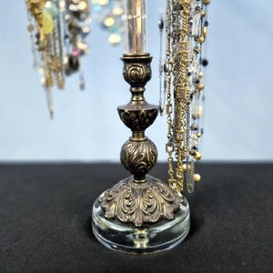 May include: A jewelry stand featuring an ornate, antique-bronze-colored metal base with intricate floral relief patterns, mounted on a clear glass circular pedestal. Multiple silver and gold-toned chains hang from the top of the central post.