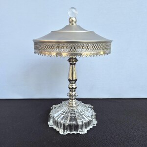 May include: A silver-toned, decorative serving dish with a clear, faceted glass base. The dish features a detailed, woven-patterned rim and a clear, spherical finial on the lid. The stem is ornate and the dish is likely for serving small items.