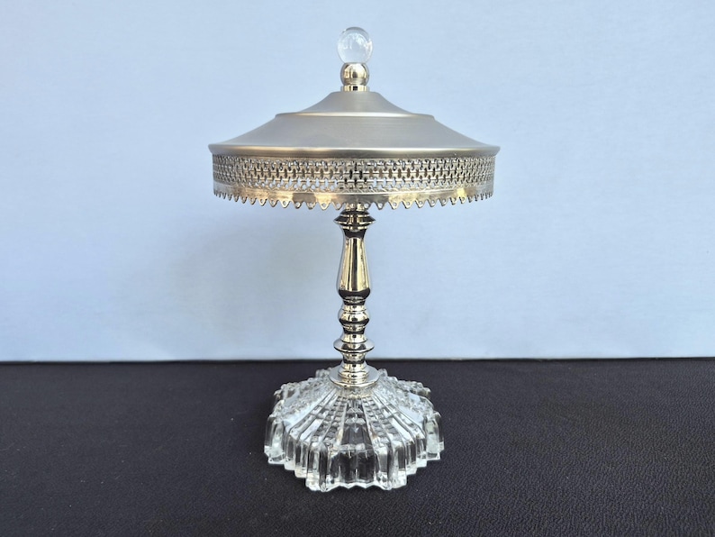 May include: A silver-toned, decorative serving dish with a clear, faceted glass base. The dish features a detailed, woven-patterned rim and a clear, spherical finial on the lid. The stem is ornate and the dish is likely for serving small items.