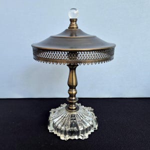 May include: A decorative stand featuring a clear, faceted glass base with a radial pattern. A central brass-toned metal pedestal supports a wide, circular brass-toned metal canopy with a delicate, perforated filigree edge and a clear glass sphere finial.