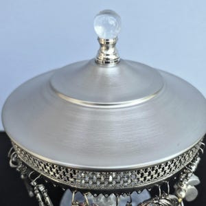 May include: A silver-toned jewelry box with a domed lid and a clear, spherical knob. The box features a decorative filigree border and dangling charms. The lid has a brushed metal finish, and the box is set against a light blue background.