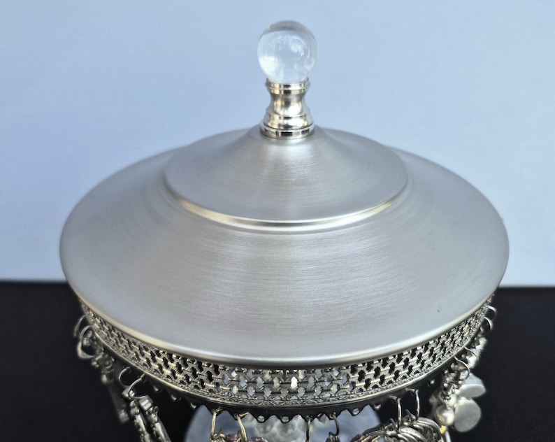 May include: A silver-toned jewelry box with a domed lid and a clear, spherical knob. The box features a decorative filigree border and dangling charms. The lid has a brushed metal finish, and the box is set against a light blue background.