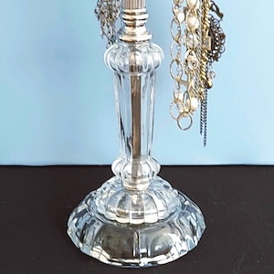 May include: A clear glass jewelry stand with a fluted pedestal base and a central column. The stand features a silver-toned metal top section with multiple hooks, currently holding various gold and silver-toned necklaces and bracelets.