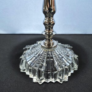 May include: A silver-colored candlestick with a clear, faceted glass base. The base has a starburst design, and the stem is a shiny, metallic silver. The candlestick is designed to hold a single candle.