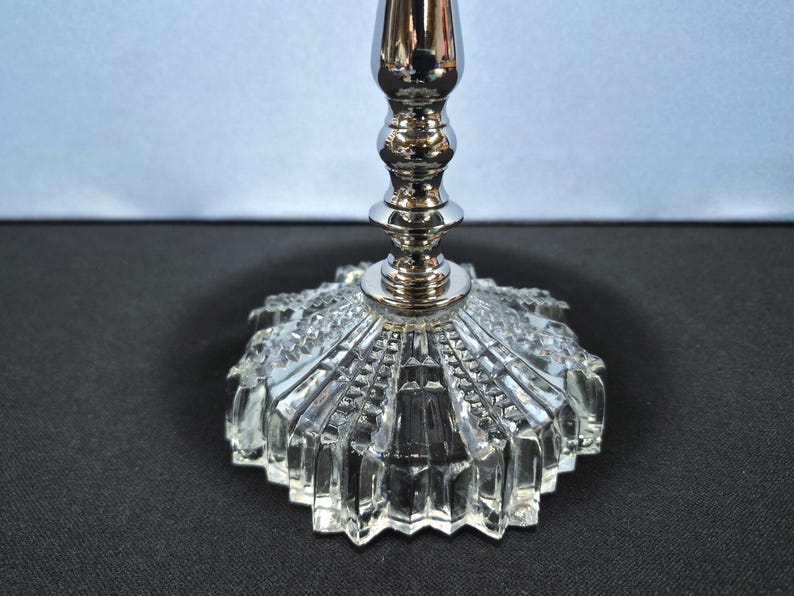 May include: A silver-colored candlestick with a clear, faceted glass base. The base has a starburst design, and the stem is a shiny, metallic silver. The candlestick is designed to hold a single candle.