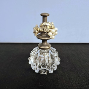 May include: A clear glass ring holder with a faceted, bulbous base and a metallic, antique-brass colored stem. The top of the stem features a flat, circular cap, and several rings with white pearls and clear stones are stacked around the post.
