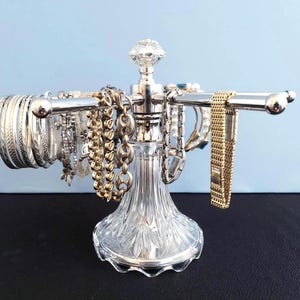 May include: A silver-toned metal jewelry display stand featuring a clear, faceted glass base and a central post topped with a decorative glass finial. The stand has horizontal arms holding various silver and gold-toned bracelets and wristwatches.