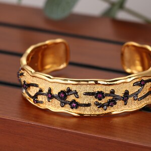 Puede incluir: Un brazalete tipo puño de tono dorado con una superficie de oro mate texturizado y un motivo de ramas de metal negro oxidado. Las ramas están adornadas con pequeñas flores de piedras preciosas de color rosa vibrante.