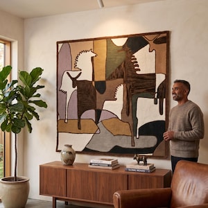 Può includere: Un grande arazzo da parete quadrato con un design geometrico astratto che presenta figure di cavalli stilizzate in tonalità marrone, crema, verde scuro e viola, incorniciato da un bordo marrone scuro e appeso a una parete chiara.