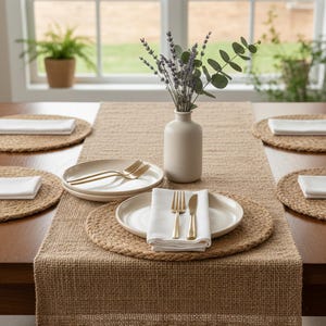 Könnte beinhalten: Ein gedeckter Tisch mit einem Tischläufer und Platzsets aus Jute. Cremefarbene Teller, goldfarbenes Besteck und weiße Servietten sind arrangiert. Eine kleine, cremefarbene Vase enthält Lavendel und Eukalyptuszweige.