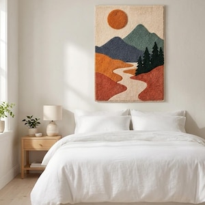 May include: A rectangular wall hanging featuring a textured punch needle landscape design. The artwork displays a burnt orange sun, blue and green mountain peaks, dark green pine trees, and a winding cream-colored river over terracotta hills.