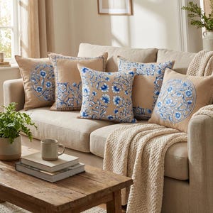 May include: A beige sofa with several decorative throw pillows. The pillows feature blue floral patterns and designs. A wooden coffee table with books and a mug is in front of the sofa. A cream-colored throw blanket is draped over the arm of the sofa.