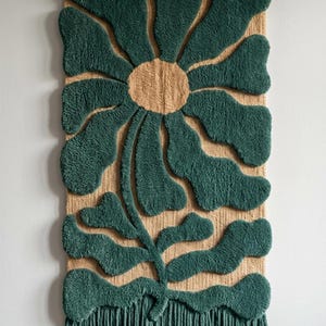 May include: A large, rectangular wall hanging featuring a stylized flower design. The flower has a tan center and dark green petals, with a matching stem and fringe. The textured, tufted design is mounted on a woven background.