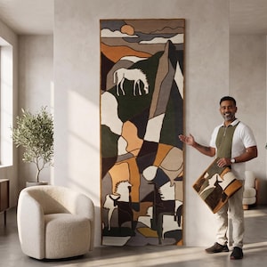 May include: A tall, rectangular wall tapestry featuring a stylized landscape with horses in shades of ochre, dark green, charcoal, and cream. The design includes abstract mountain shapes and a sun. A matching square cushion is held in the foreground.