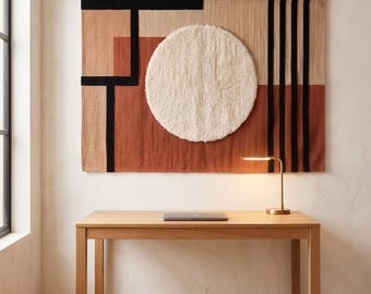 May include: A rectangular wall hanging featuring a geometric composition of black, tan, and terracotta woven fabric with a large, central, circular off-white tufted element. The piece is displayed above a light-colored wooden desk with a lamp.