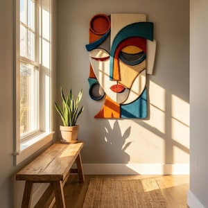 Może przedstawiać: Prostokątna dekoracja ścienna przedstawiająca stylizowaną, abstrakcyjną twarz, złożoną z warstwowych, teksturowanych fragmentów tkaniny w żywych odcieniach turkusu, pomarańczu, czerwieni, żółci i kremu, z wyraźnymi czarnymi konturami.