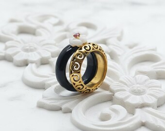 May include: Two rings, one black and one gold, are stacked together. The gold ring has an openwork design. A small white flower with a pink center stone sits atop the rings. The rings are displayed on a white, floral-patterned surface.