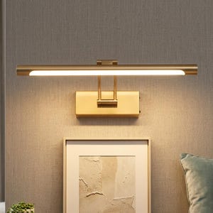 May include: A horizontal wall-mounted picture light with a brushed brass finish. The fixture features a long, cylindrical lamp head with an integrated LED light strip that emits a warm glow, connected to a rectangular wall plate by two arms.