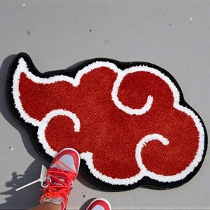 May include: Red cloud-shaped rug with white swirls and a black border. The rug is on a gray surface. A person's red and white sneakers are visible near the rug.