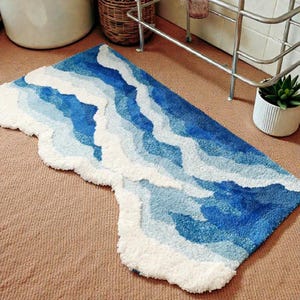 May include: A rectangular bath mat with a wavy design in shades of blue and white, resembling ocean waves. The mat has a soft, textured surface and a unique, irregular edge. The rug is placed on a brown carpet.
