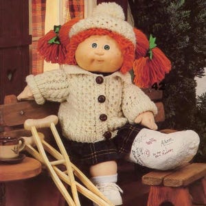 Puede incluir: Muñeca Cabbage Patch Kid con coletas de hilo rojo y un gorro y suéter de punto color crema. La muñeca lleva una falda a cuadros, zapatos blancos y una escayola en una pierna. Un par de muletas de madera y una mesa pequeña con una taza están en primer plano.