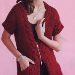 May include: A burgundy cable-knit vest with short sleeves and a full-length zip. The vest has two front pockets and a hood. The model is wearing a silver bracelet and a necklace with a pendant. The background is a solid pink.