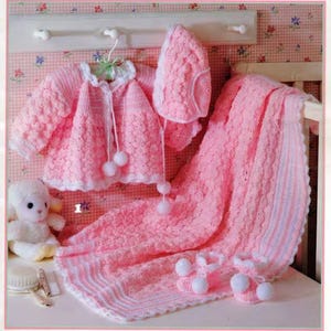 May include: A baby's crochet set in soft pink, including a jacket, hat, blanket, and booties. The jacket and hat have white trim and pom-poms. The blanket and booties have a textured pattern. A small stuffed lamb and grooming tools are also visible.
