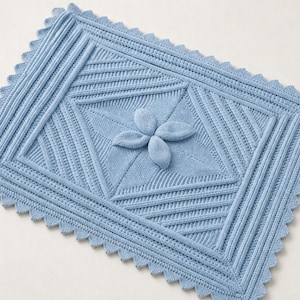 May include: A light blue knitted blanket with a central flower design. The blanket features a geometric pattern with a scalloped edge. The flower is made of four rounded petals. The blanket is approximately 76 cm long and 51 cm wide.