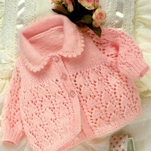 May include: A light pink knitted baby cardigan with a scalloped collar and sleeves. The cardigan features a delicate leaf pattern on the front and is fastened with three clear, decorative buttons. The garment is displayed on a white surface with pink flowers.