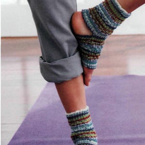 May include: A person stretches on a purple yoga mat, wearing grey trousers and colourful knitted foot warmers. The foot warmers have a striped pattern in shades of blue, green, red, and white. The person's foot is flexed.