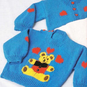 Op de afbeelding: Twee blauwe gebreide truien. Eén is een ronde hals met een teddybeerontwerp en rode harten. De andere is een vest met hartvormige knopen en rode hartaccenten. Beide truien hebben geribbelde manchetten.