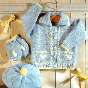 May include: A baby blue knitted set comprising a jacket, hat, and mittens. The jacket features yellow trim, buttons, and pom-poms. The set is displayed against a wooden backdrop with a yellow ribbon.