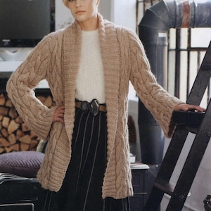 Op de afbeelding: Een lange, beige kabelgebreide cardigan met een sjaalkraag. De cardigan wordt gedragen over een witte trui en een zwart-wit gestreepte rok. Een bruine riem met een decoratieve gesp accentueert de taille.