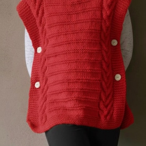 Op de afbeelding: Een rode gebreide poncho met een coltrui en drie grote witte knopen. De poncho heeft een kabelgebreid ontwerp aan de zijkanten en horizontale ribbels aan de voorkant. Het kledingstuk wordt gedragen over een wit shirt en een zwarte broek.