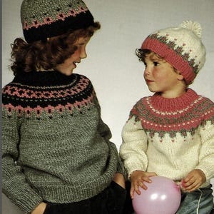 May include: Two children wearing matching knitted sweaters and hats. The sweaters are gray and cream-colored, with a patterned yoke in pink, black, and gray. The hats have a matching design and a pom-pom on top. One child holds a pink balloon.