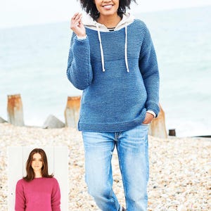 May include: A woman wearing a blue knit hoodie with a white drawstring and a light-colored hood. She is also wearing light blue jeans. The background is a beach with wooden posts and the ocean.