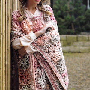 May include: A handmade crochet shawl in shades of pink, beige, and gray. The shawl features a floral pattern and a fringed edge. The garment is worn over a light-colored shirt and is suitable for cool weather.