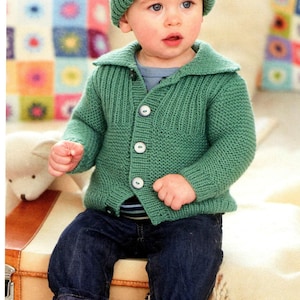 Puede incluir: Un bebé con gorro y cárdigan verde tejido, con botones blancos, y vaqueros azul oscuro. El niño está sentado en una maleta vintage, con un peluche y una manta cerca.