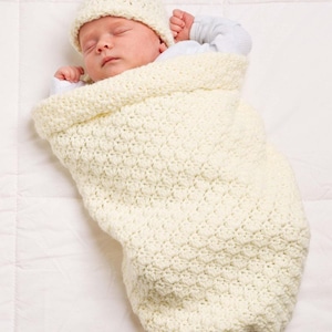 Op de afbeelding: Een slapende baby gewikkeld in een crèmekleurige, gehaakte cocon en bijpassende muts. Het getextureerde breiwerk biedt warmte en comfort. De baby draagt een lichtblauwe onesie. De achtergrond is een wit, gewatteerd oppervlak.
