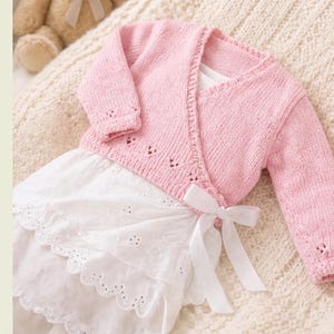 May include: A light pink knitted wrap cardigan with long sleeves and a delicate eyelet pattern along the cuffs and hem, layered over a white sleeveless dress featuring a scalloped eyelet lace skirt and a white ribbon tie at the waist.
